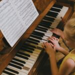 A young girl with blond hair playing piano, following music sheets indoors.
